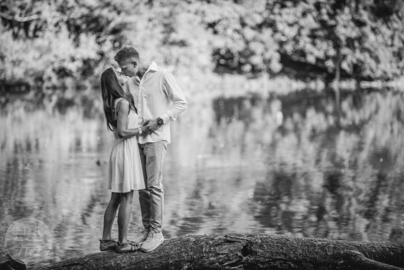 Czarno-białe zdjęcie pary całującej się na powalonym pniu drzewa nad wodą, z odbiciem drzew w tle.