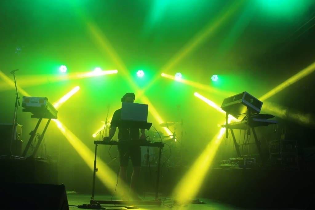Artysta w słuchawkach, grający na syntezatorze z laptopem na scenie oświetlonej zielonymi i żółtymi reflektorami.