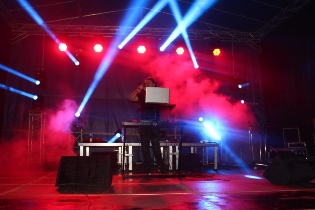 DJ na scenie oświetlonej czerwonymi i niebieskimi reflektorami, z laptopem i efektami dymnymi.