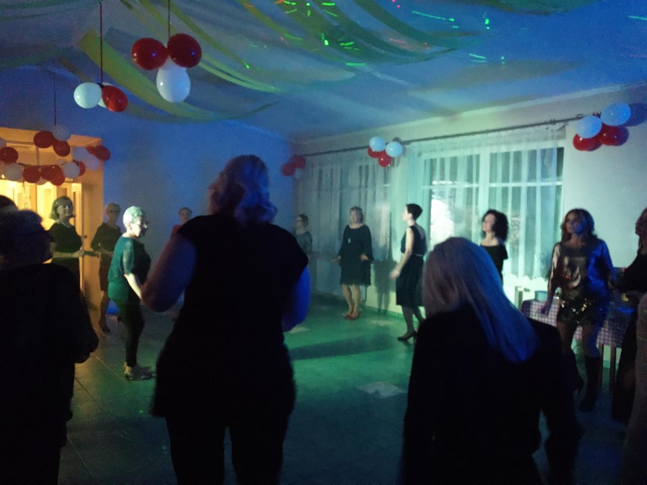 Sala balowa z dekoracjami balonowymi i serpentynami, oświetlona zielonym i niebieskim światłem, z grupą kobiet w różnym wieku w ciemnych strojach.