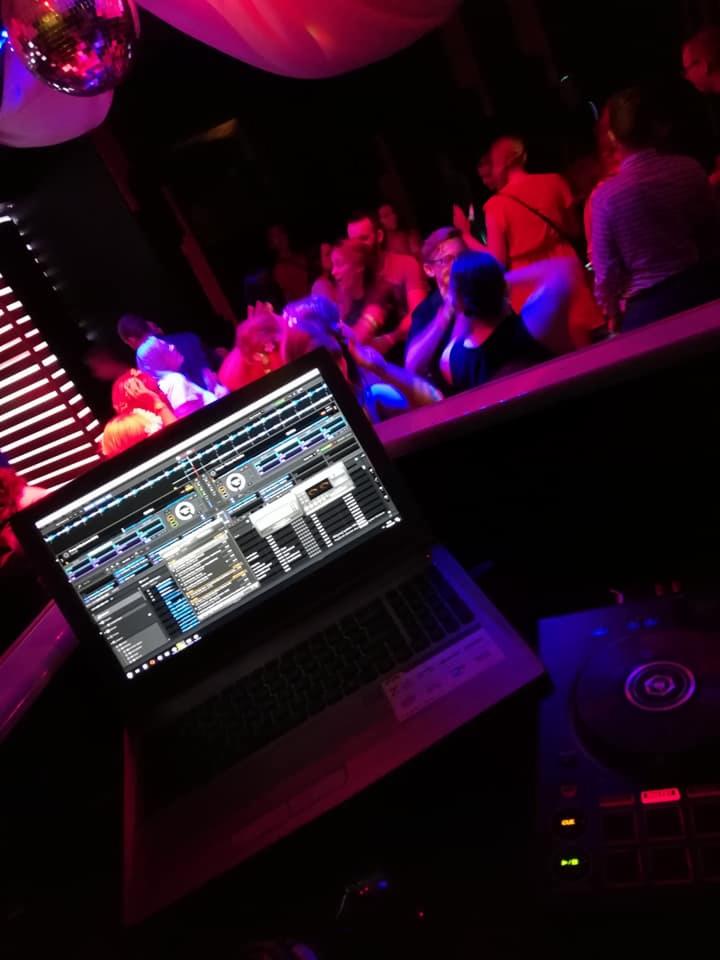 Laptop z oprogramowaniem DJ-skim na tle roztańczonego tłumu w klubie, oświetlonym kolorowymi światłami, obok mikser DJ-ski.