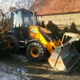 Żółto-czarna koparko-ładowarka JCB z łyżką na nierównym terenie, w tle budynek z czerwoną dachówką i częściowo ośnieżona kostka brukowa.