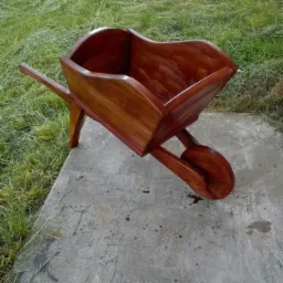 Dekoracyjna, mała taczka z ciemnego drewna, stojąca na betonowej płycie obok trawnika.