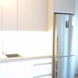 Minimalistyczna zabudowa kuchenna z górnymi szafkami w odcieniach beżu, białymi dolnymi szafkami i stalową lodówką, całość oświetlona ledami.