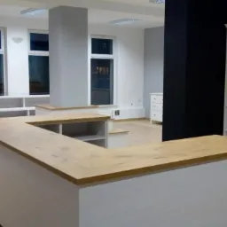 Nowoczesne wnętrze biurowe z recepcją o blacie drewnianym, widok na białe regały i filary w jasnym pomieszczeniu z oknami.