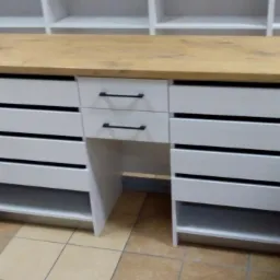 Biała szafka na wymiar z szufladami z poziomymi szczelinami i blatem z jasnego drewna, widok z przodu na tle płytek podłogowych i regałów.