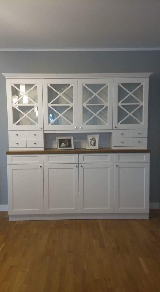 Biała, klasyczna witryna z przeszklonymi drzwiczkami z krzyżowym wzorem, drewniany blat, zdjęcia w ramkach, jasna podłoga.