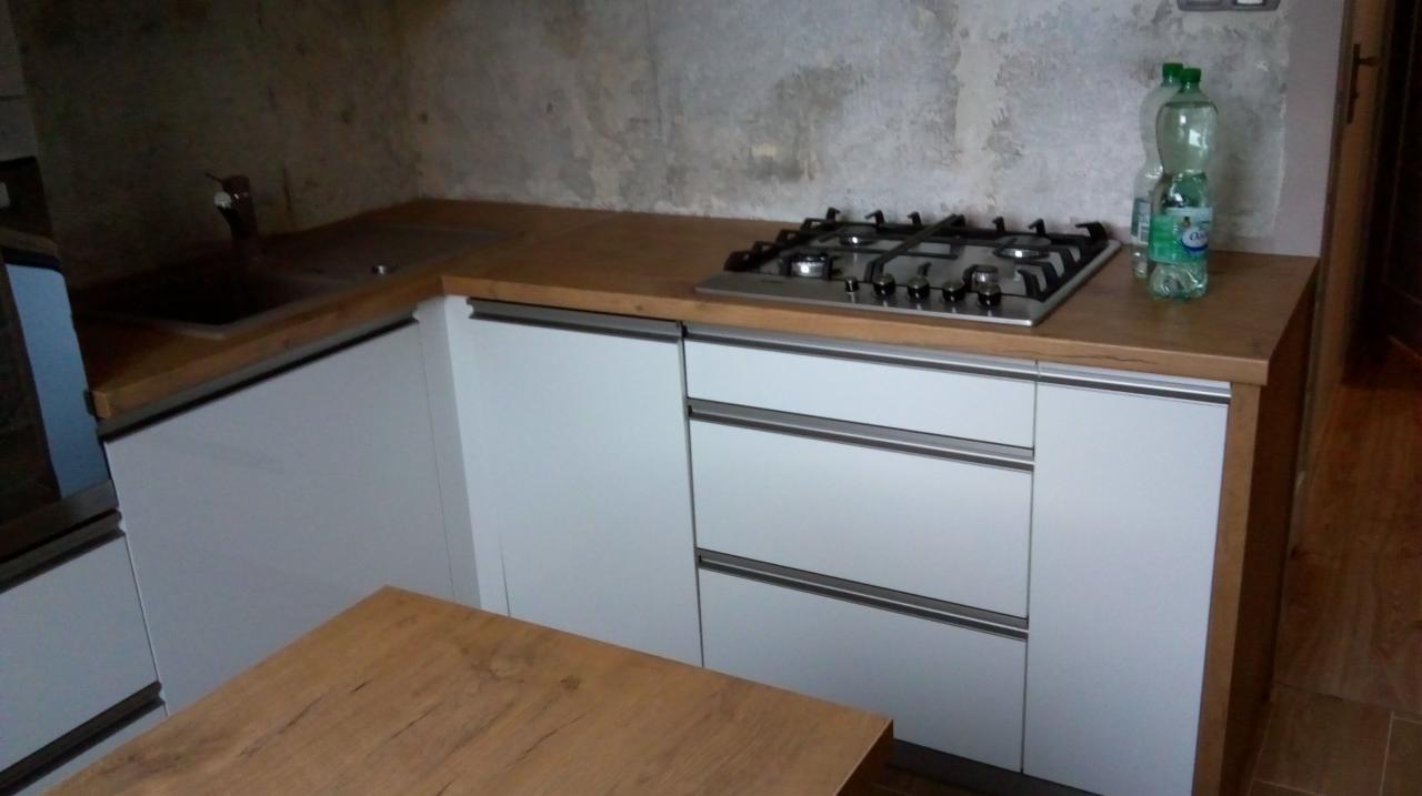 Fragment kuchni z białymi szafkami, drewnianym blatem, płytą gazową i butelkami wody na tle surowej ściany.