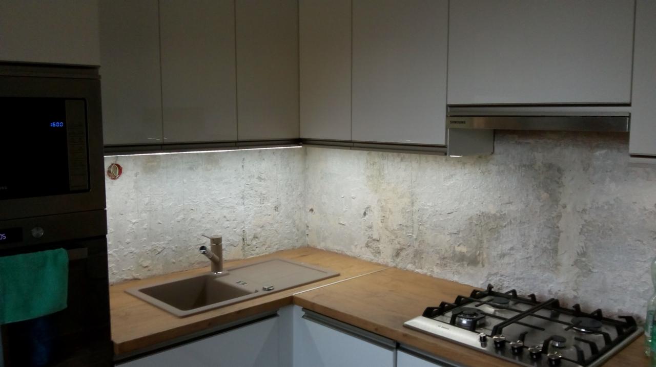 Fragment kuchni z drewnianym blatem, beżowym zlewem i kuchenką gazową, oświetlony ledami, na tle ściany o surowym wykończeniu przypominającym beton.