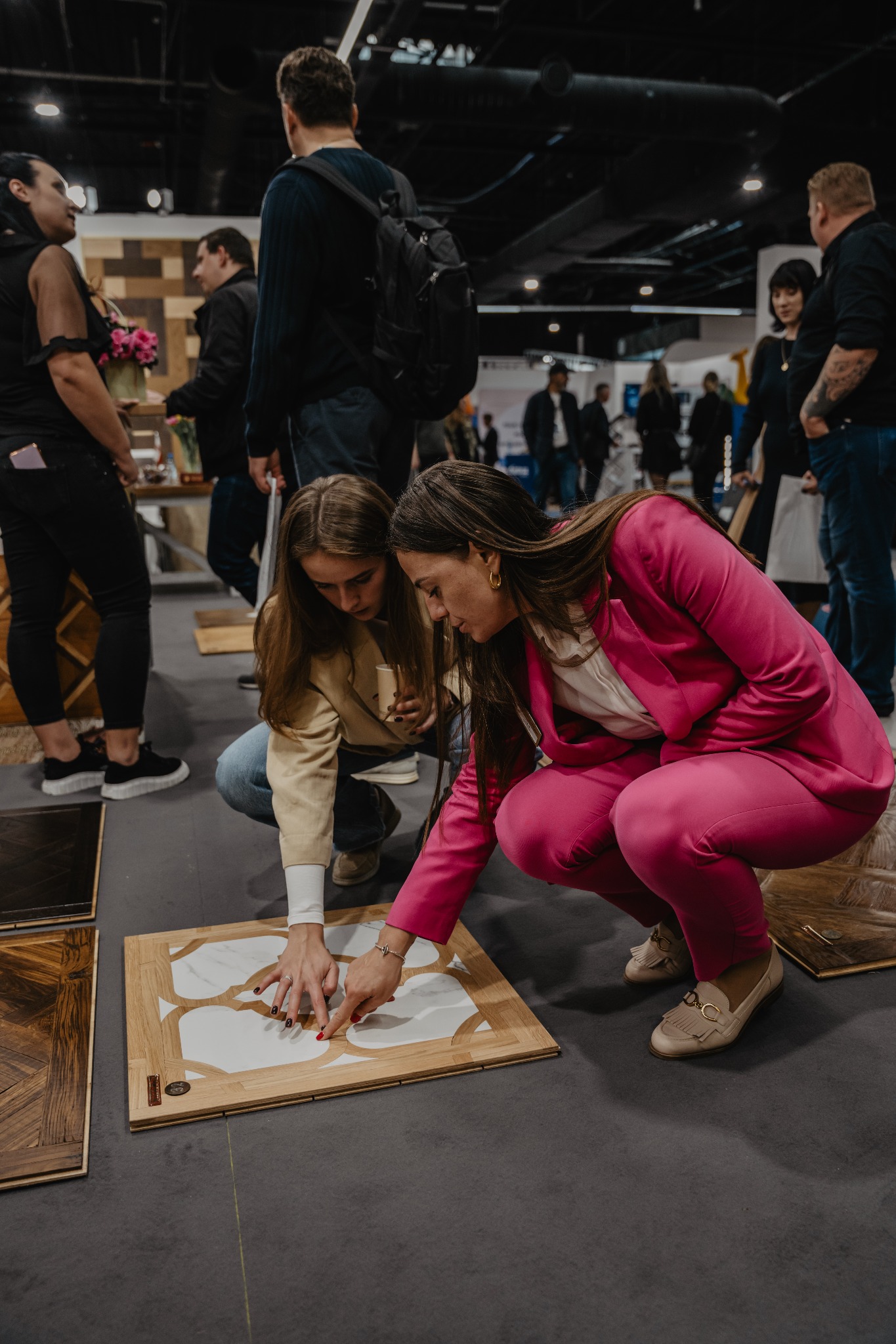 Dwie kobiety pochylone nad próbką podłogi z drewnianą ramą i białym wzorem marmuru, prezentowaną na targach w otoczeniu innych próbek i zwiedzających.
