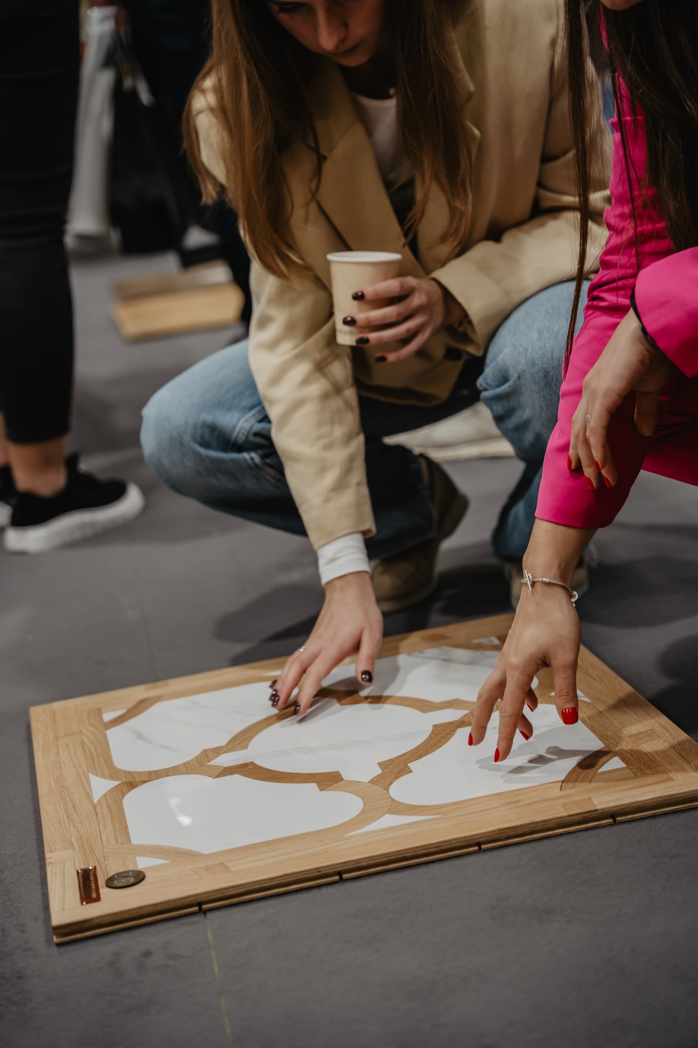Dwie kobiety pochylone nad próbką parkietu z drewnianą ramą i białym, wzorzystym wypełnieniem, jedna trzyma kubek z kawą, a druga ma na nadgarstku srebrną bransoletkę.