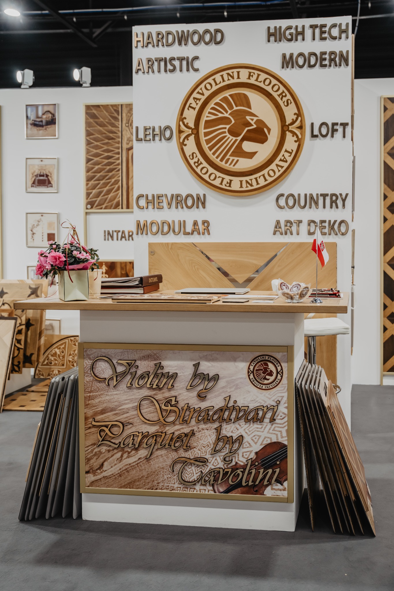Ekspozycja producenta parkietów Tavolini Floors na targach, prezentująca wzorniki parkietów, logo firmy z lwem, hasła reklamowe oraz aranżację z kwiatami i flagą Polski.