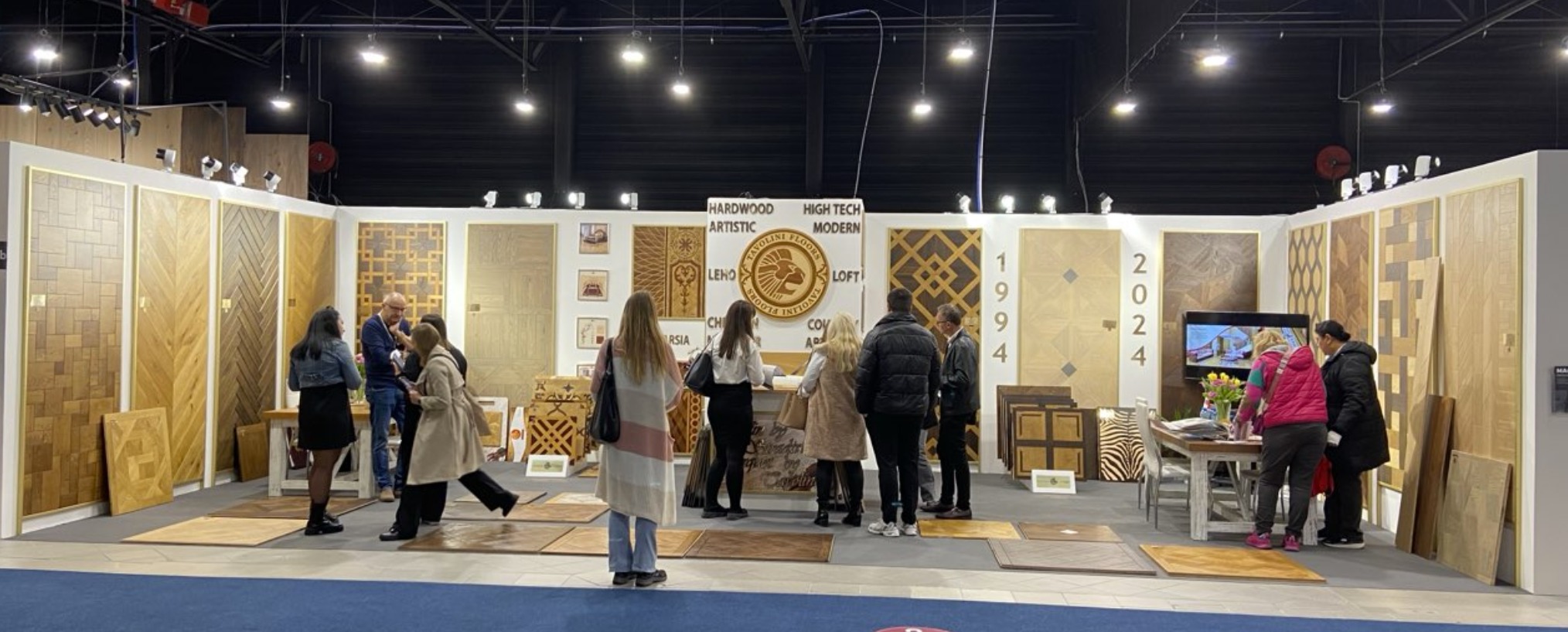 Ekspozycja różnorodnych wzorów parkietów i paneli drewnianych na targach, z odwiedzającymi oglądającymi próbki i rozmawiającymi przy stoisku firmy Tavolini Floors.