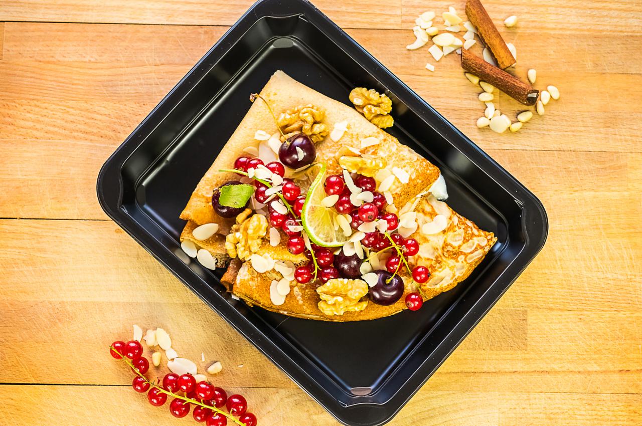 Dwa naleśniki na czarnym talerzu, udekorowane czerwoną porzeczką, wiśniami, orzechami włoskimi, płatkami migdałów i plasterkiem limonki, obok laska cynamonu i gałązka porzeczki na drewnianym tle.