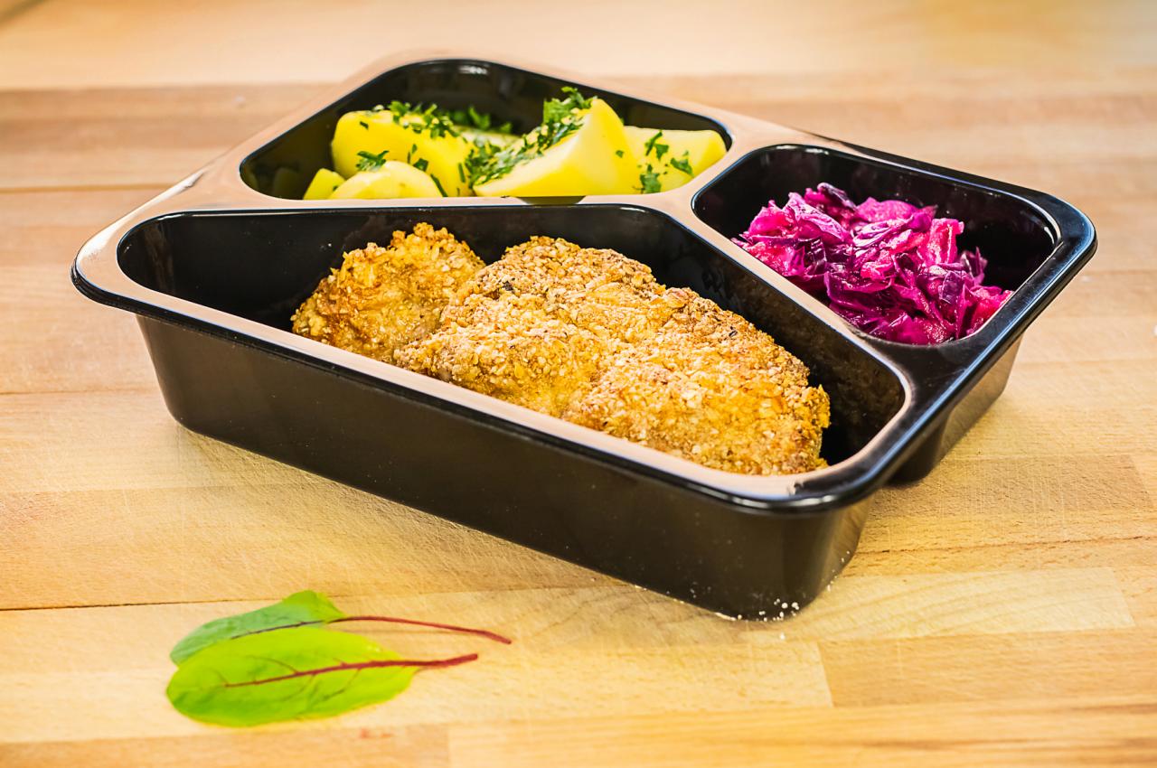 Gotowe danie w czarnym, trójdzielnym pojemniku: kotlety w panierce z sezamu, ziemniaki z koperkiem i czerwona kapusta, całość na drewnianym blacie z dwoma listkami sałaty.