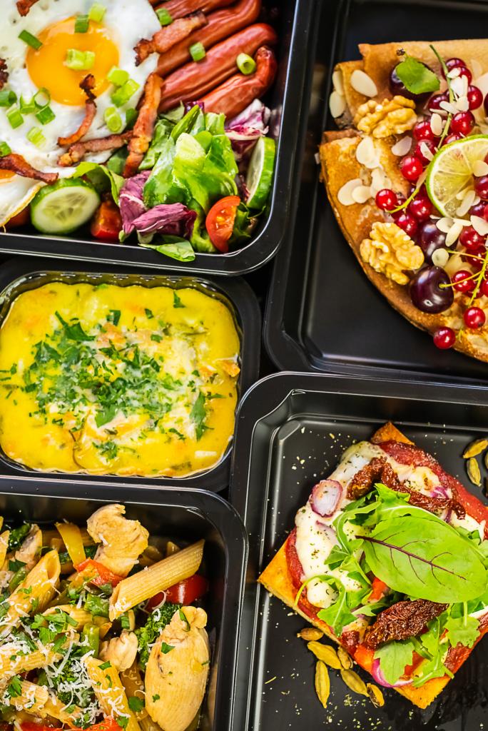 Zestaw pięciu dań w czarnych pojemnikach: śniadanie z jajkiem sadzonym i bekonem, kremowa zupa, makaron z kurczakiem i warzywami, naleśnik z owocami i orzechami, oraz pizza z suszonymi pomidorami...