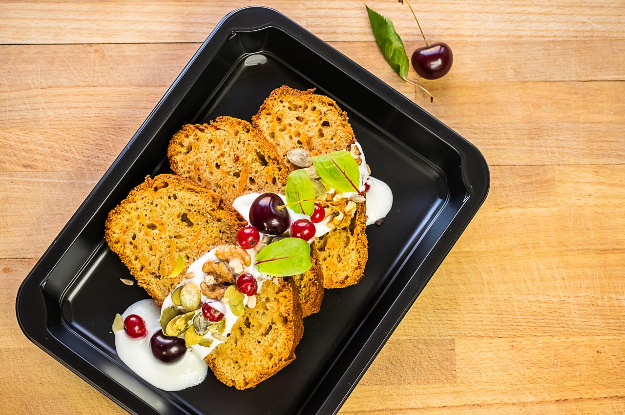 Kawałki ciasta marchewkowego na czarnej blasze, polane białym sosem i udekorowane wiśniami, orzechami włoskimi i listkami, z pojedynczą wiśnią obok blachy na drewnianym tle.