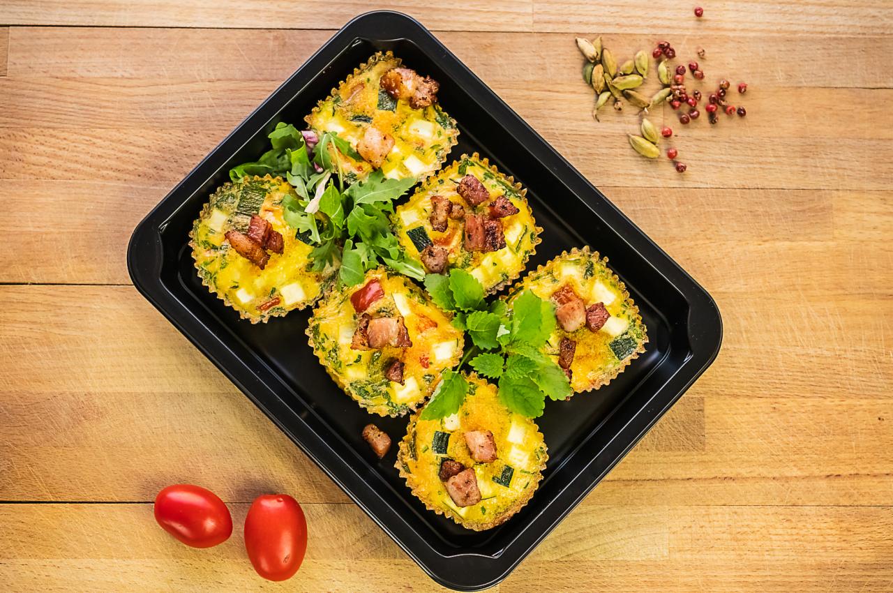 Sześć wytrawnych babeczek z jajkiem, warzywami i boczkiem ułożonych w czarnej foremce na drewnianym stole, udekorowanych świeżymi ziołami, z dodatkiem pomidorków koktajlowych i przypraw.