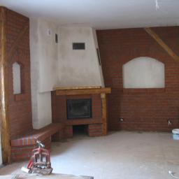 Kominki Szablinski - Wnętrze w trakcie budowy z ceglaną obudową paleniska z drewnianą belką nad otworem, widoczny murarz i narzędzia.