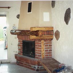 Kominki Szablinski - Murowany piec z drewnianymi belkami, czarnym wkładem i dekoracyjnymi kamieniami na ścianie, widok z boku na otwarte drzwi balkonowe.