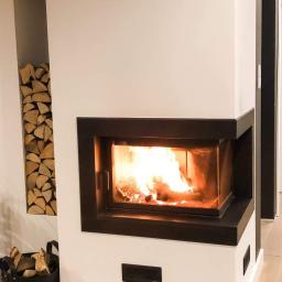 Kominki Szablinski - Wnętrze z nowoczesnym, narożnym paleniskiem z czarną obudową, płomienie tańczą za szybą, drewno ułożone w niszy obok, kosz z drewnem w pobliżu.