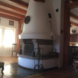 Kominki Szablinski - Duży, wolnostojący piec kaflowy z kamienną obudową i metalowymi drzwiczkami, w przestronnym salonie z drewnianymi belkami stropowymi i widoczną kuchnią w tle.