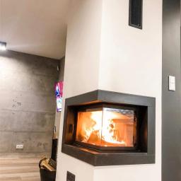 Kominki Szablinski - Nowoczesny narożny wkład kominkowy z przeszklonym frontem, płomienie widoczne wewnątrz, obudowa w kolorach bieli i grafitu, drewno do palenia w wiaderku obok.