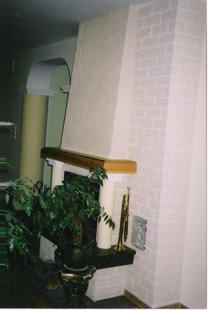 Wykończony kominek z obudową z jasnej cegły, drewnianym blatem i dekoracyjną rośliną w ozdobnej donicy. Z prawej strony trąbka i płaskorzeźba.