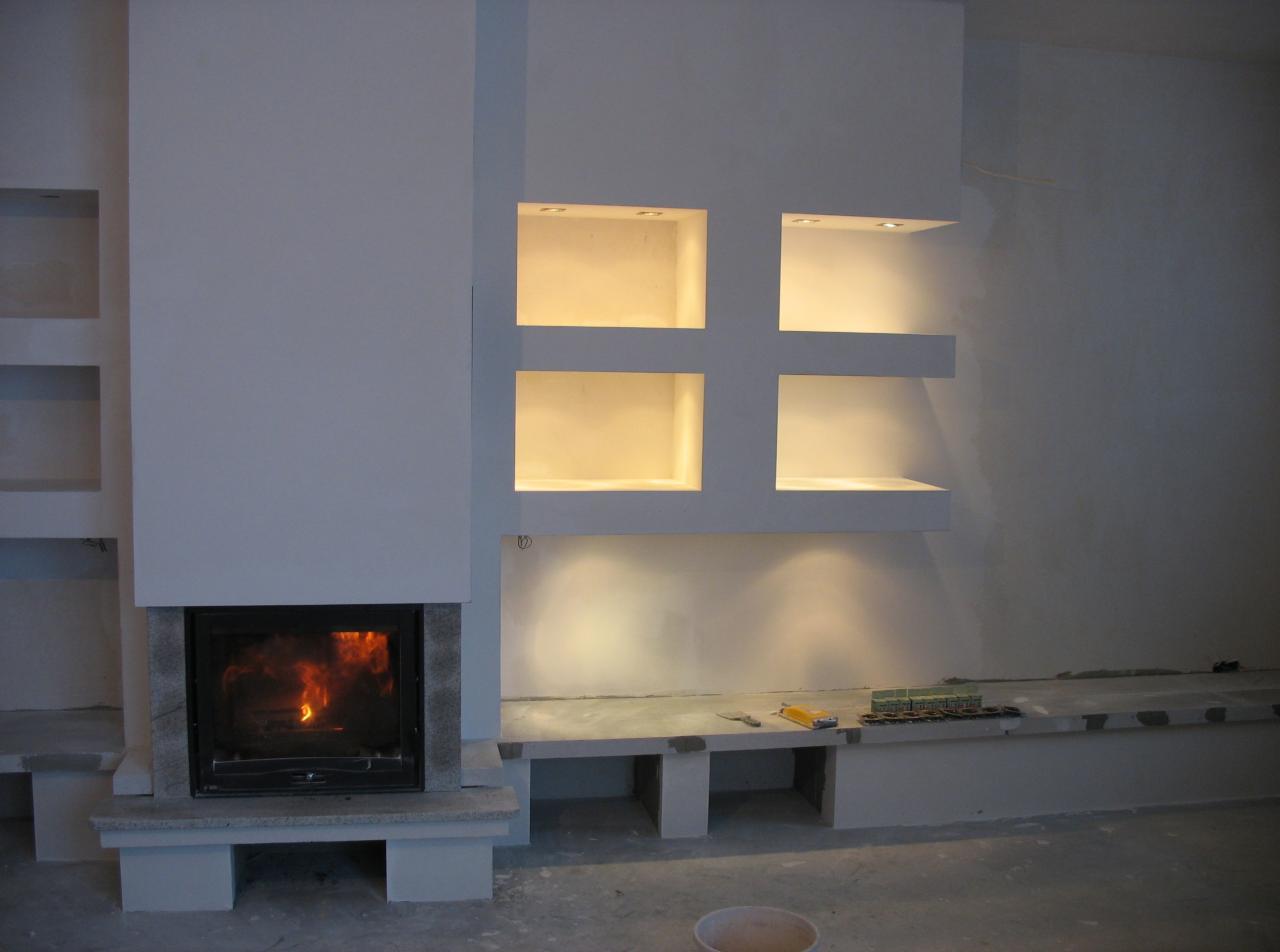 Nowoczesny wkład kominkowy z paleniskiem obudowany jasnym kamieniem, zintegrowany z minimalistyczną ścianą z oświetlonymi niszami, widoczne narzędzia i materiały budowlane sugerują trwający montaż.