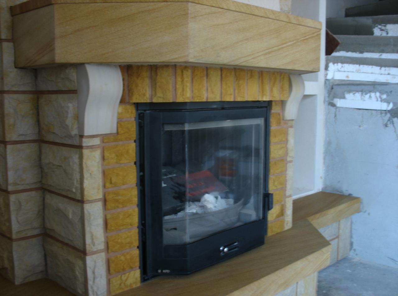 Zamknięty wkład kominkowy obudowany piaskowcem w odcieniach beżu i żółci, z widocznymi fragmentami surowych schodów w tle.