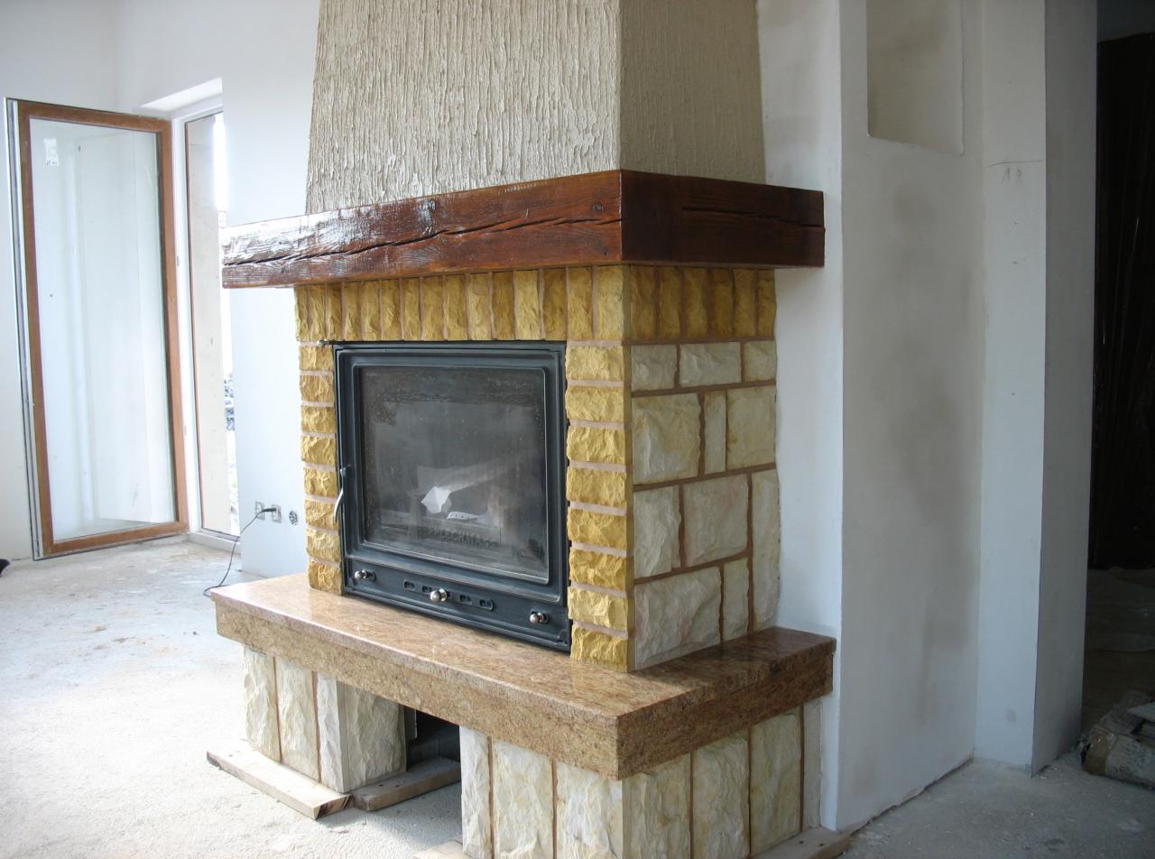 Murowany wkład kominkowy z obudową z piaskowca i drewnianą belką nad paleniskiem, w surowym wnętrzu z oknem.