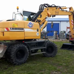 Żółta koparka kołowa Liebherr 314 Litronic z łyżką i lemieszem, zaparkowana na trawiastym terenie, z widocznym logo Poltrans i butlami gazowymi w tle.