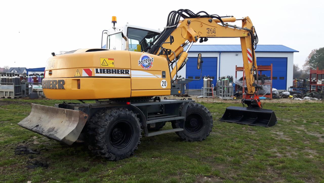 Żółta koparka kołowa Liebherr 314 Litronic z łyżką i lemieszem, zaparkowana na trawiastym terenie, z widocznym logo Poltrans i butlami gazowymi w tle.