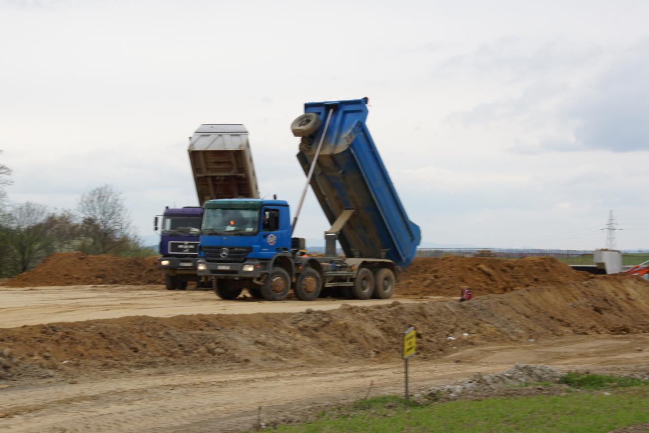 Dwa wywrotki, niebieski Mercedes i fioletowy MAN, rozładowują ziemię na placu budowy drogi w Nysie, widoczne kopce ziemi i elementy konstrukcji drogi w tle.