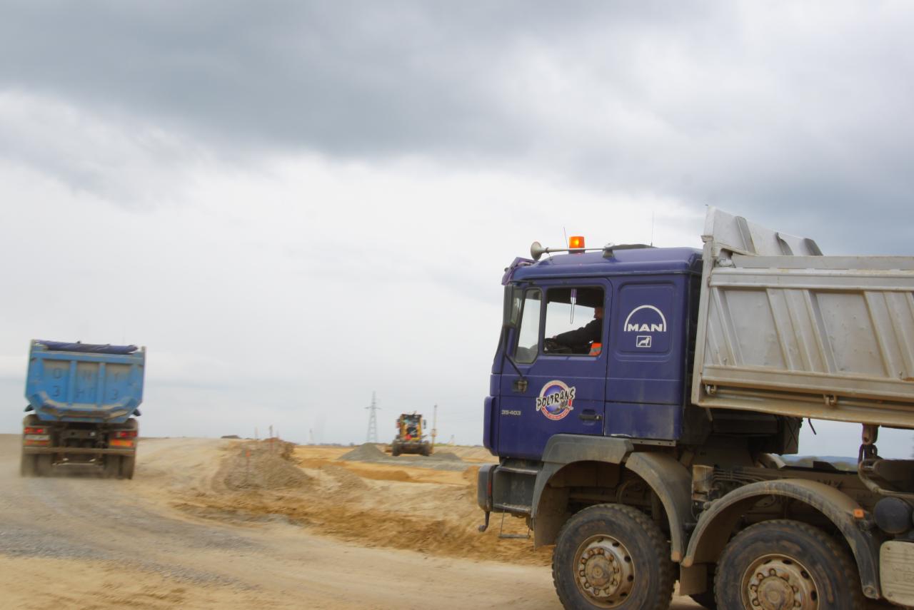 Ciężarówka MAN na placu budowy z innymi pojazdami w tle, pochmurne niebo.