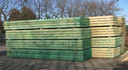 Stosy impregnowanego drewna budowlanego, zabezpieczonego sznurami, na placu składowym, widoczne drzewa w tle.