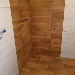 Ukończona kabina prysznicowa z drewnopodobnymi płytkami na ścianach i podłodze, oraz białymi płytkami imitującymi marmur. Widoczne elementy instalacji wodnej.