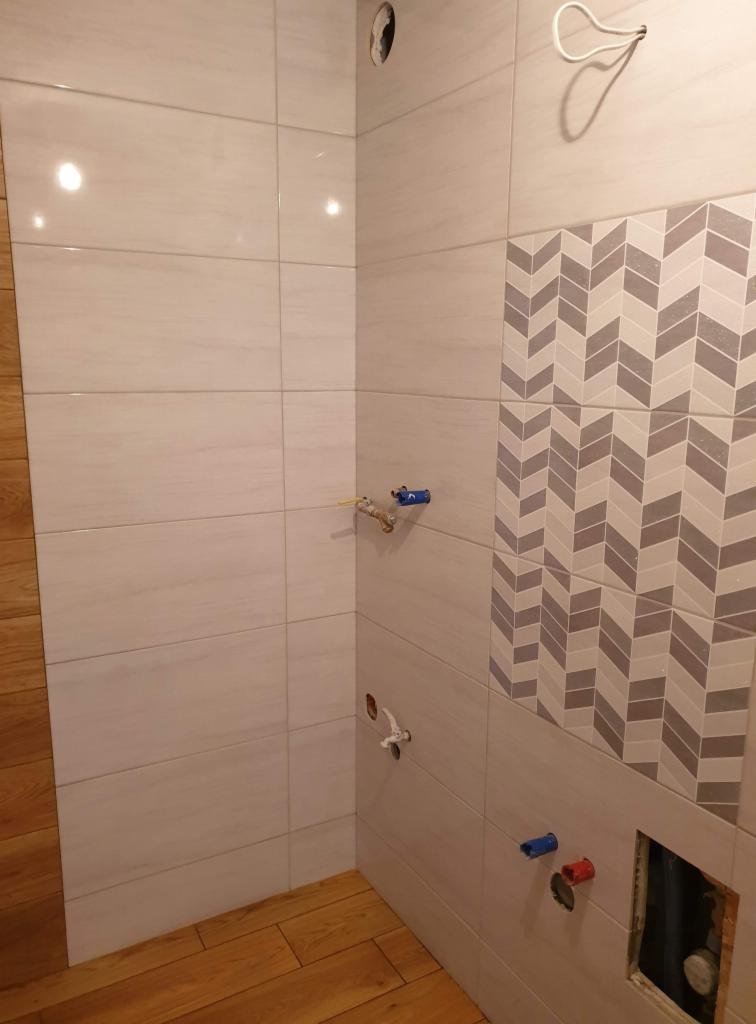Fragment wykończonej łazienki z jasnymi płytkami imitującymi drewno, dekoracyjnym pasem z wzorem chevron i wystającymi elementami instalacji wodnej.