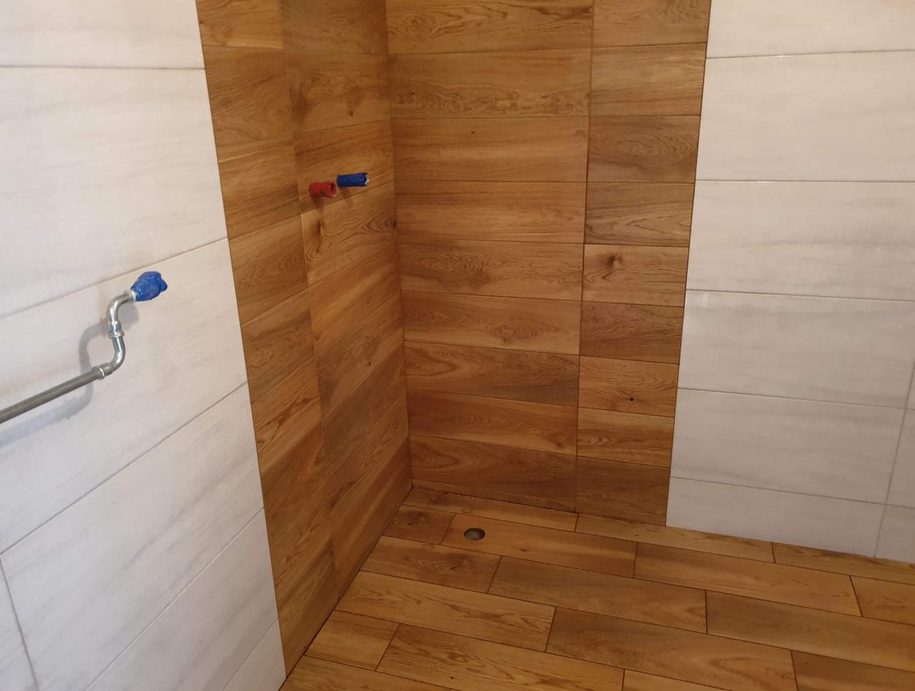 Ukończona kabina prysznicowa z drewnopodobnymi płytkami na ścianach i podłodze, oraz białymi płytkami imitującymi marmur. Widoczne elementy instalacji wodnej.