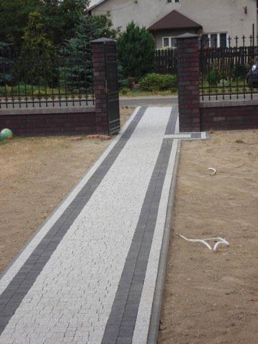 Ułożona ścieżka z jasnoszarej kostki brukowej z ciemnoszarymi obrzeżami prowadząca do bramy z ceglanymi słupkami na tle domu i zieleni.