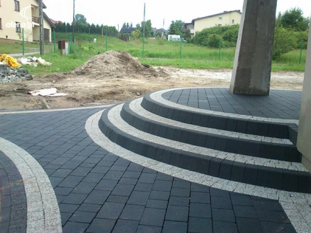 Schody z ciemnej kostki brukowej z jasnymi obramowaniami, prowadzące do betonowego filaru, w tle widoczny plac budowy i domy.