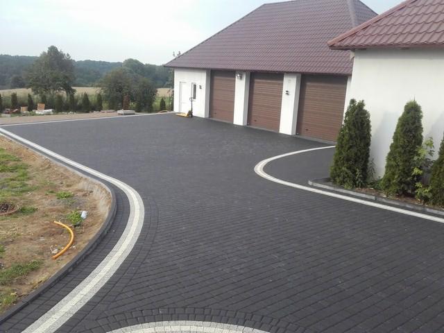 Czarna kostka brukowa ułożona na podjeździe do garażu z trzema bramami, obrzeża wykończone białymi pasami, widok z góry na posesję.