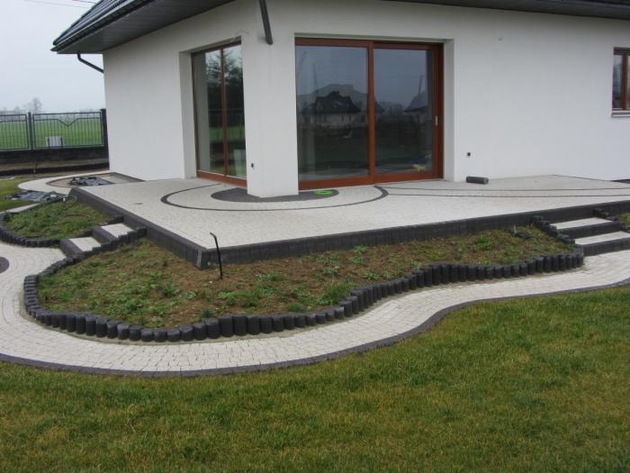 Wykończony taras z jasnej kostki brukowej z ciemnymi obramowaniami, schodkami i rabatami, przylegający do białego domu z brązowymi oknami przesuwnymi.