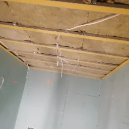 Montaż izolacji z wełny mineralnej w suficie podwieszanym z płyt kartonowo-gipsowych, widoczne profile metalowe i przewody elektryczne.