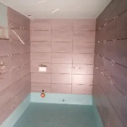 Łazienka w trakcie remontu: położone płytki ścienne w odcieniach brązu z krzyżykami dystansowymi, hydroizolacja podłogi w kolorze turkusowym, wystające przewody elektryczne z sufitu z płyty karton...