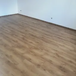 Ukończone podłogi z paneli laminowanych w jasnym odcieniu dębu, widoczne listwy przypodłogowe i przygotowane otwory na gniazdka elektryczne.