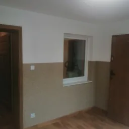 Pokój po remoncie z drewnopodobną podłogą, ściany z dolną częścią pokrytą beżową strukturą, okno z widoczną drabiną w tle i brązowe drzwi.