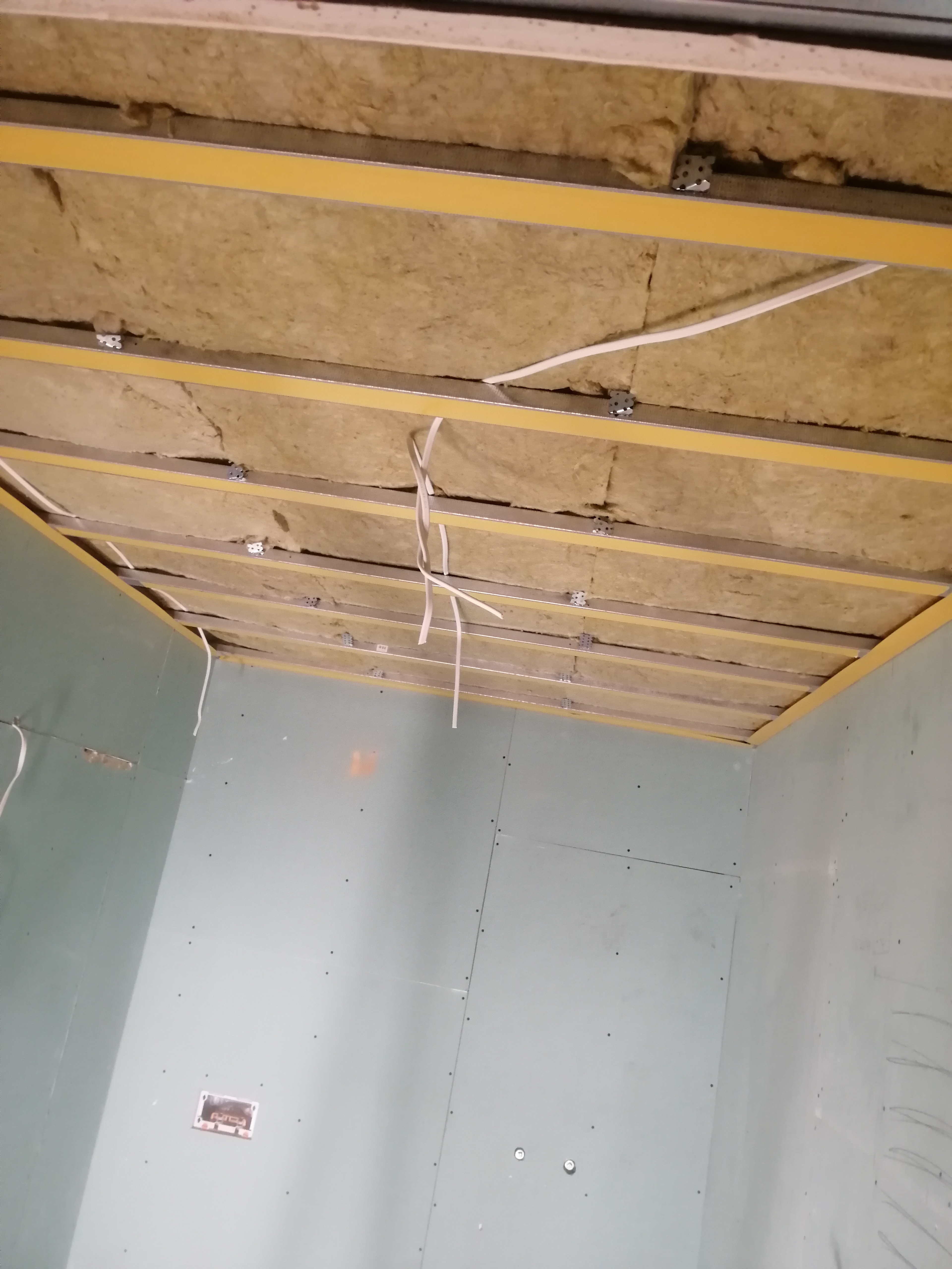 Montaż izolacji z wełny mineralnej w suficie podwieszanym z płyt kartonowo-gipsowych, widoczne profile metalowe i przewody elektryczne.