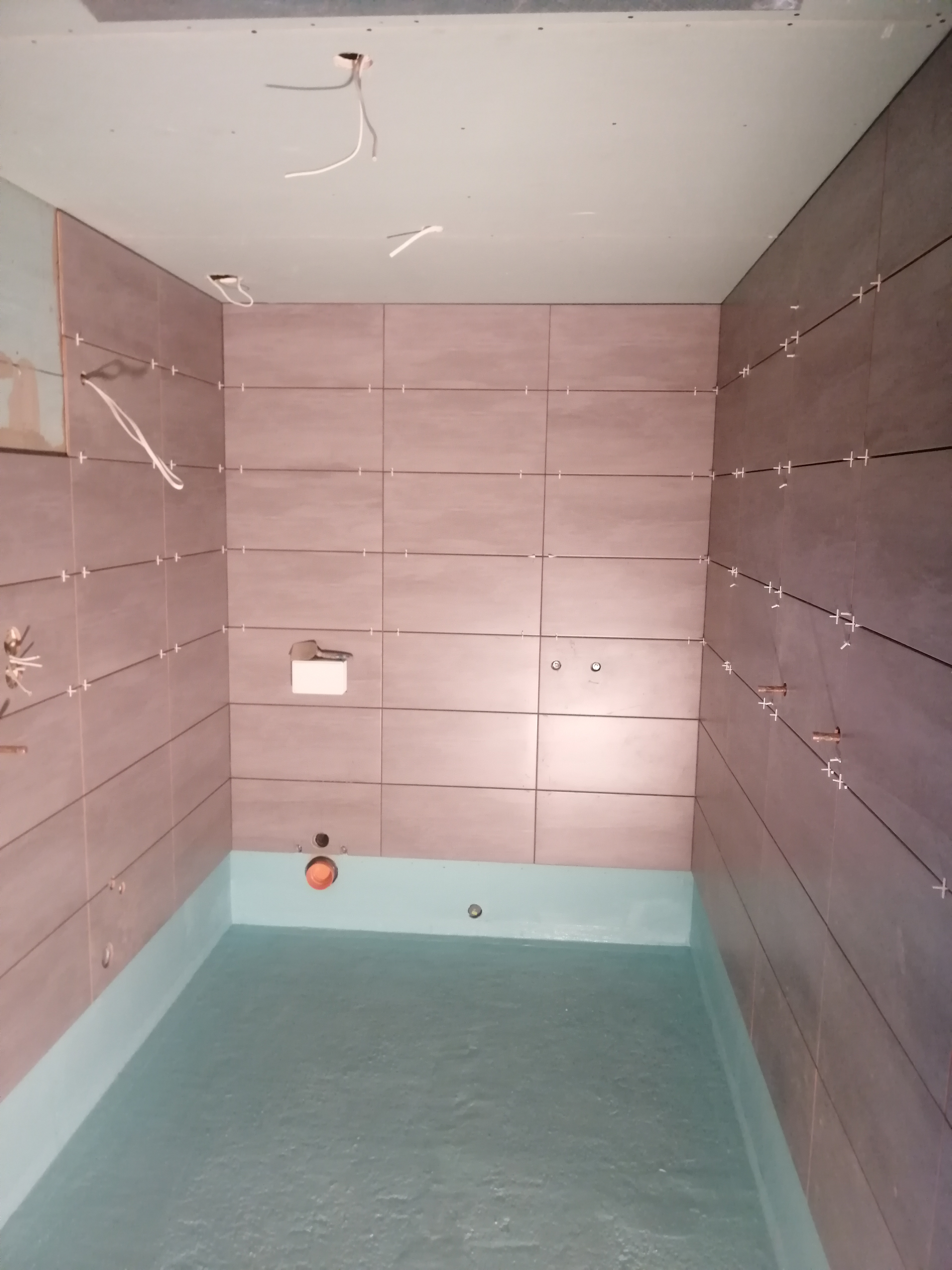 Łazienka w trakcie remontu: położone płytki ścienne w odcieniach brązu z krzyżykami dystansowymi, hydroizolacja podłogi w kolorze turkusowym, wystające przewody elektryczne z sufitu z płyty karton...