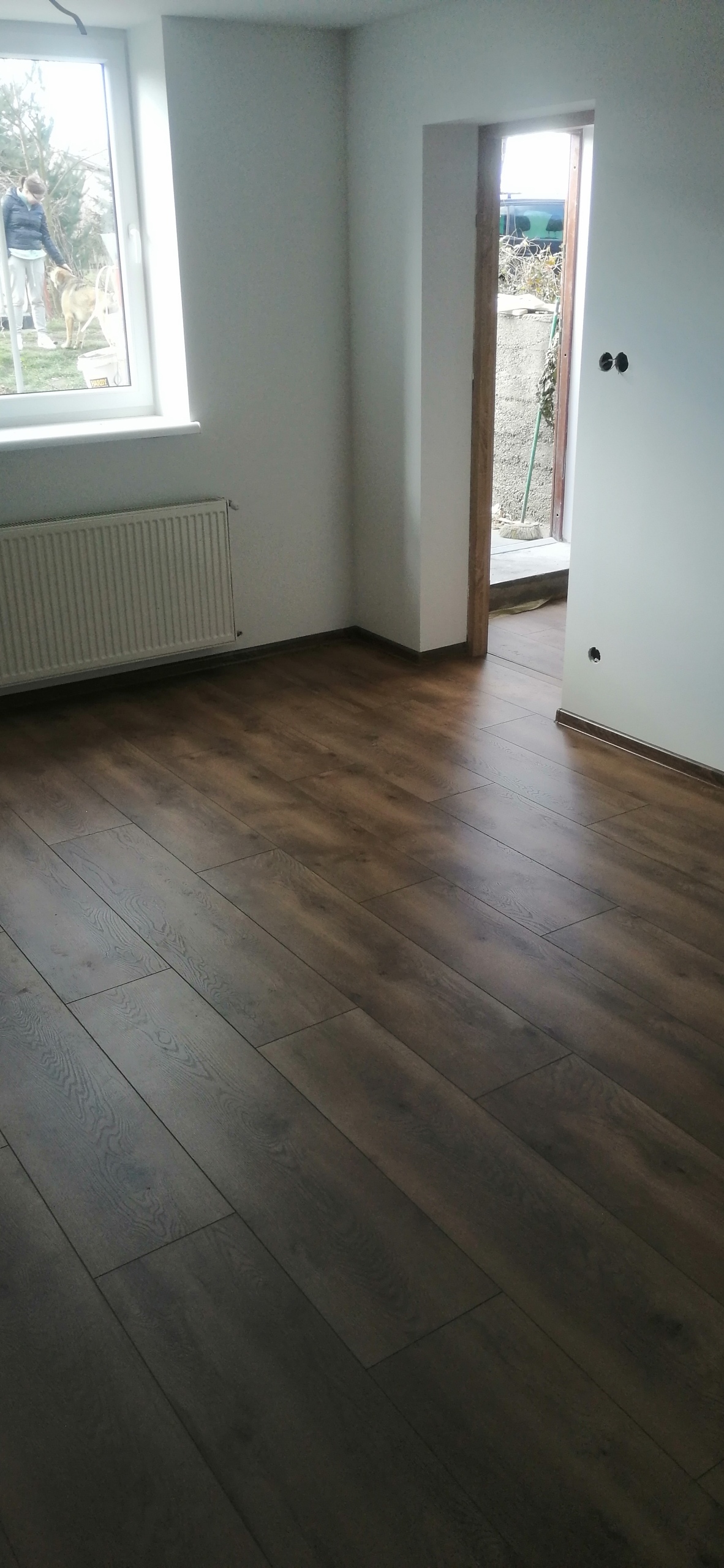 Ułożone ciemne panele podłogowe w pustym, świeżo wyremontowanym pokoju z grzejnikiem i widokiem na zewnątrz przez okno, gdzie widać osobę z psem.